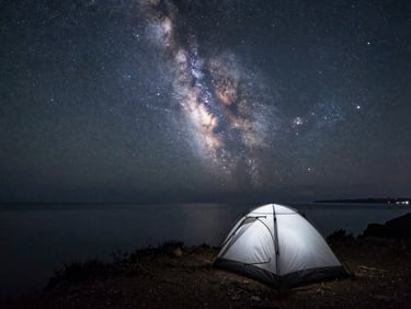 Wide starscape over a single silhouetted tent pitched on a coastal cliff, the Milky Way glowing softly above a calm dark sea.