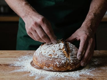 Close-up of a baker's hands dusting flour over a freshly proofed sourdough loaf on a wooden peel, soft directional light, deep shadows, matte finish, 35mm film grain.