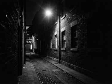 Gritty black and white 35mm film shot of a dark London alleyway at night under a single harsh street lamp, deep shadows, high contrast, heavy film grain.