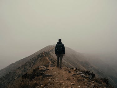 Wide environmental shot of a lone traveler standing on a misty mountain ridge, viewed from behind, overcast lighting, desaturated earth tones, 35mm cinematic photography