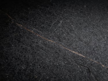 Tight macro shot of a dark, textured graphite stone surface with subtle bronze veins, dramatic side-lighting, high contrast, architectural, 35mm
