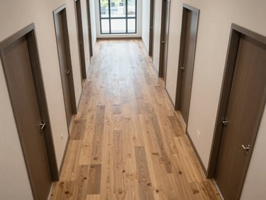 Overhead view of a multi-family residential corridor with fresh LVP flooring installed, unit doors lining both sides, overcast natural light from end windows, no occupants, clean material edges visible