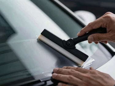 Close-up of a technician's hands smoothing ceramic film onto a car window with a squeegee, tight macro shot, high-contrast lighting, deep charcoal and cool white tones.