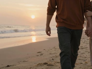 An intimate close-up of a couple walking along a foggy beach at sunset, warm low-angle light reflecting on the sand, holding hands gently.