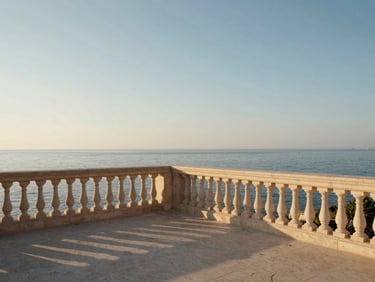 Wide architectural shot of a Mediterranean coastal terrace at golden hour, stone balustrade in the foreground, calm sea filling the far distance, warm raking light across pale stone, no people, generous sky