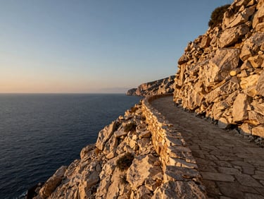 Wide landscape of a Mediterranean coastal cliff at golden hour, stone path curving toward the horizon at the right third, sea stretching to the left, no figures present, warm low-angle light on rock face