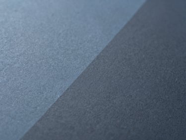 Extreme close-up of fine textured paper with abstract geometric shadows, minimalist editorial photography, soft diffused daylight, muted stellar blue and charcoal tones, 35mm