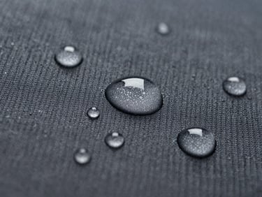 A macro detail shot of water droplets perfectly beading on a textured dark gray ripstop fabric surface, bright cleanroom daylight, extreme close-up.