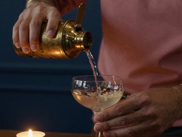 Close-up of hands pouring liquid from a brass shaker into a coupe glass, warm incandescent light, deep shadows.