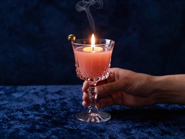 Hands holding a warm, smoking cocktail in a heavy crystal glass, low-lit candle light, dark blue velvet background.