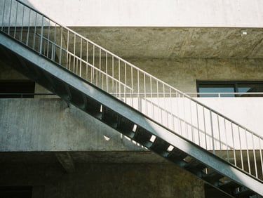Architectural shot looking up toward a concrete mezzanine and minimalist steel staircase, sharp daylight streaming from above, 35mm lens.