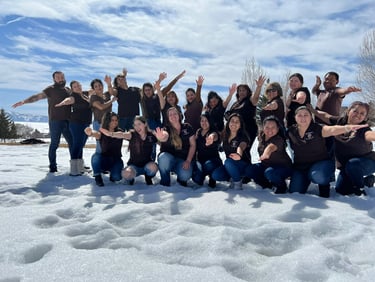A diverse group of coworkers in brown shirts posing and waving in the snow during a winter team building event.