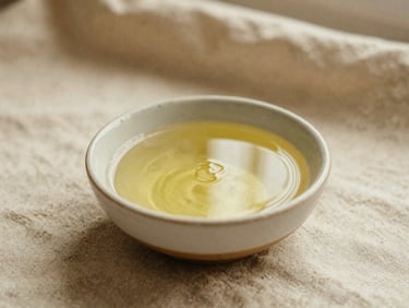 Warm organic oil in a small ceramic bowl, soft natural linen background, warm sand tones, editorial photography