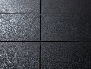 Straight-on macro shot of rubber gym flooring tiles, interlocking seam visible at centre, dense black surface texture under clean overhead studio light, no people, geometric precision
