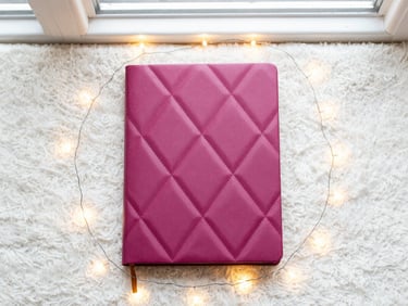 Cozy flatlay of a journal with a diamond pattern cover, surrounded by sparkling fairy lights, on a soft white rug, warm and inviting