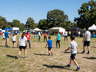 Wide environmental view of an outdoor community cultural festival on a sunny day, families gathered, active sports stations in the background, bright daylight, 35mm lens