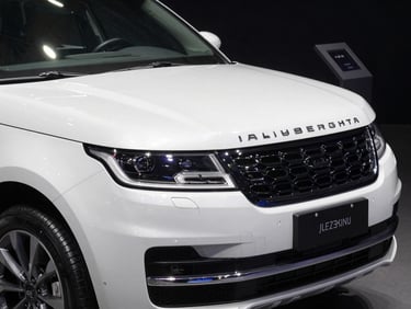 Tight three-quarter front detail of a white luxury SUV on a showroom floor, dramatic overhead strobe lighting creating sharp reflections along the hood and grille, near-black background, no people visible