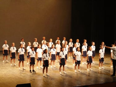 Wide environmental shot, a large group of school-age dancers in a full formation on a lit concert stage, warm golden stage spotlights, Vinod visible at the edge of the frame gesturing direction, the formation filling the full frame, candid live-performance energy