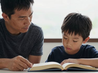 Close framing of a parent and child seated across from each other at a table, both looking down at the same open book, overcast daylight from the right, attention and presence emphasized, no eye contact or smiling, quiet focused interaction, high contrast