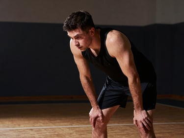 Tight portrait of an athlete in a training hall, head slightly bowed, eyes locked on a fixed point ahead, hands resting on knees, studio strobe lighting from the upper left casting sharp shadows, dark background, solitary figure, no teammates