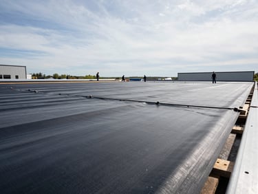 Low-angle wide shot of a large commercial flat roof under active installation, crew members visible across the span, seaming membrane in daylight, overcast New England sky, sharp material detail in foreground
