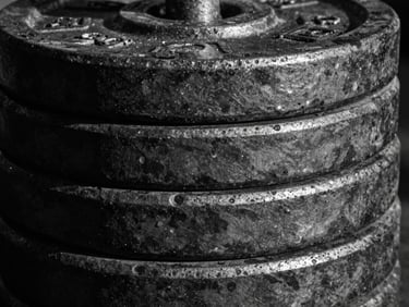 Extreme close-up of a stack of heavy iron weight plates, black and white, high contrast, textured metal, with harsh side lighting creating deep shadows.