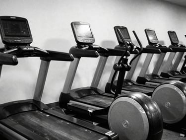 Empty cardio row with treadmills and elliptical machines, black and white, high contrast, reflections on screens, clean and spacious feel, harsh side lighting.