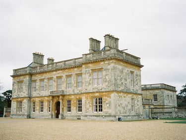 Wide architectural angle of a classic stone English manor estate under soft overcast British daylight, low-contrast natural lighting, desaturated slate and champagne tones, 35mm film.