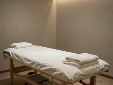 Wide environmental shot of a clean minimal treatment room in warm diffused light, a massage table centered in frame with neatly folded linen, pale warm-gray walls, no clutter, subtle teal accent in towel detail on the left edge