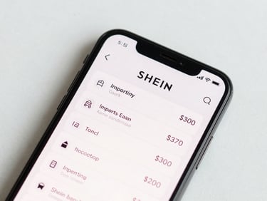 Close-up overhead flat-lay of a smartphone screen showing the Shein app order summary with import tax line visible, clean studio daylight, white background, sharp detail
