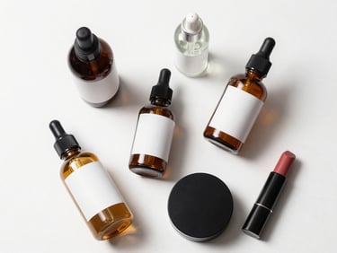 Close overhead flatlay of beauty products — skincare bottles, a compact, and a lip product arranged on a white surface with soft studio light, labels not readable, clean minimal styling