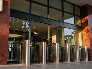 Low-angle architectural shot of a full-height steel turnstile integrated into a modern corporate lobby, warm golden-hour daylight streaming through glass, soft motion blur of a person passing through.