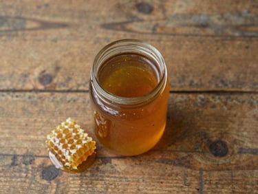 Overhead flat-lay of a single glass jar of honey on a worn wooden table, soft diffused daylight, one honeycomb shard beside it, no other objects in frame