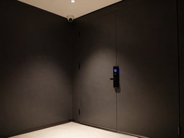 Wide-angle corner shot of a modern entry hall at night, a digital door lock with a faint blue LED ring mounted flush on a dark wall, a compact security camera angled downward in the upper frame corner, precise architectural lighting, no people
