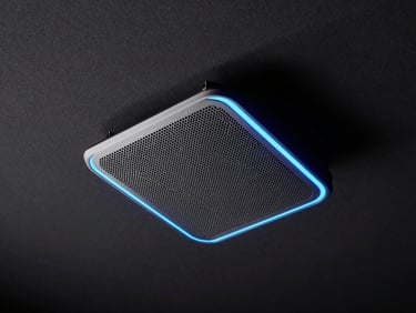 Dramatic low-angle shot of a ceiling-flush in-wall speaker installed in a dark home theater ceiling, a neon-blue edge light tracing the speaker grille perimeter, near-black surround, studio strobe catching the metal mesh texture