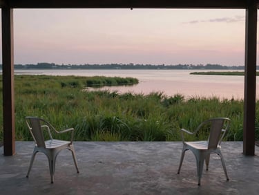 Wide environmental shot of a lagoon-edge room's exterior terrace at dusk — simple iron chairs facing open water, soft green marsh grasses in the middle distance, the lagoon surface reflecting fading pink sky, no people, overcast diffuse light
