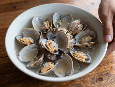 Overhead close-up of a plain ceramic bowl of clams in broth on a rough wooden table, a hand reaching in from the right edge, natural diffuse window light, no styling props, documentary food photography