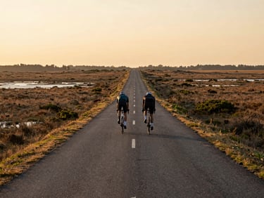 Telephoto shot of two cyclists at distance on a long straight delta path, flat wetland landscape stretching to the horizon, golden-hour side light, no posing or eye contact