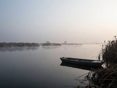 Wide panoramic Po Delta lagoon in deep winter, thick mist sitting over still dark water, silhouette of a single flat-bottomed boat moored at a reed-bank edge on the right third, overcast diffuse pale light, absolute stillness, no human figures visible