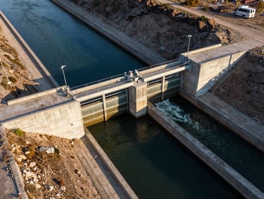Aerial view of a completed concrete weir and canal diversion structure, surrounding terrain visible, clear water flowing through controlled gates, late afternoon light casting long shadows