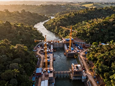 Aerial view of a hydroelectric construction site in dense Brazilian terrain, heavy cranes and formwork visible along a river bend, equipment operators working at scale, golden-hour daylight raking across the valley