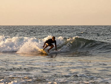 Surfer mid-paddle in shallow broken whitewater, shot from the shoreline at golden hour, wide frame showing the full arc of the bay behind, water catching low orange light, no staged pose