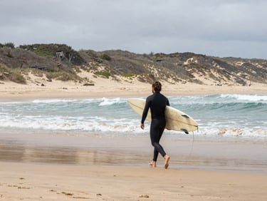 Overcast afternoon at Pipa beach, a surfer walking toward the waterline carrying a board under one arm, dune backdrop, ocean in the middle distance, no pose, movement captured mid-stride, wide framing showing beach environment