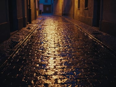 Cinematic 16:9 still of wet cobblestones reflecting warm amber streetlights in a narrow alley, deep shadows, rich navy tones, 35mm grain