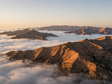 Wide panoramic aerial view of mountain ranges at golden hour, soft clouds below, warm sunlight hitting the peaks, fine art photography style