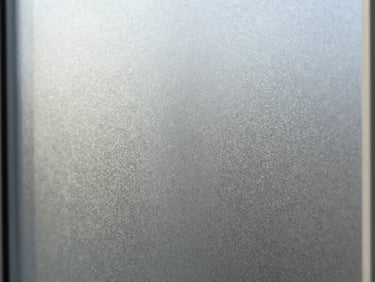 Close-up of a frosted glass panel surface under natural window light, showing even satin texture and diffused light transmission, 1/4-inch thickness, edge sharp and clean