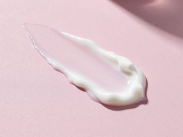 Close-up of a creamy white cosmetic lotion swatch on a dusty pink background, soft shadows, natural morning light, clean editorial texture