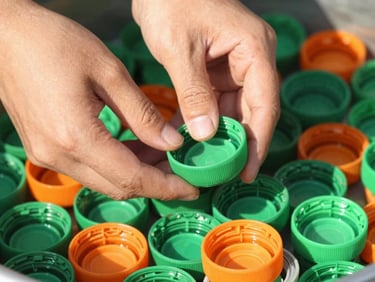 Close-up of hands sorting vibrant green and orange plastic bottle caps under warm morning sunlight, shallow depth of field.