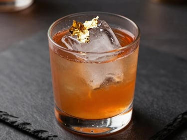 A vibrant signature cocktail with clear ice block, decorated with edible gold leaf, placed on dark slate. Warm ambient lighting, luxury editorial style.