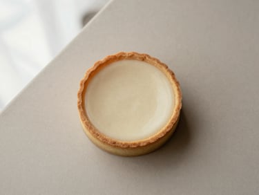 Overhead flat lay of a minimalist individual pastry, possibly a tart or entremet, with clean lines and a subtle texture, on a light earth-toned surface. Soft, directional natural light.